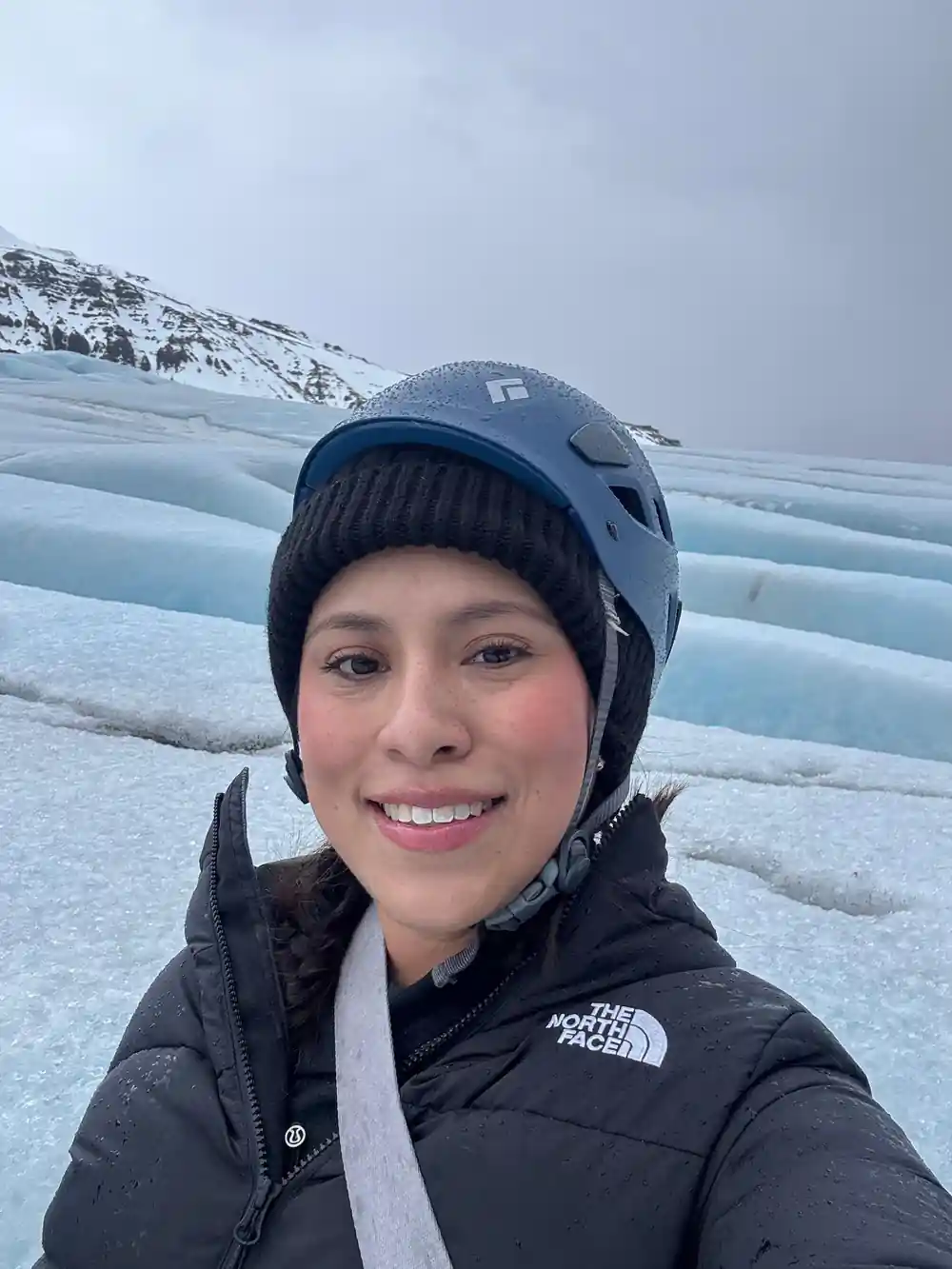 Woman in helmet smiling at glacier with mountains behind